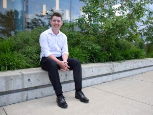 A young man sits on a ledge in front of a building with a raised garden behind him.