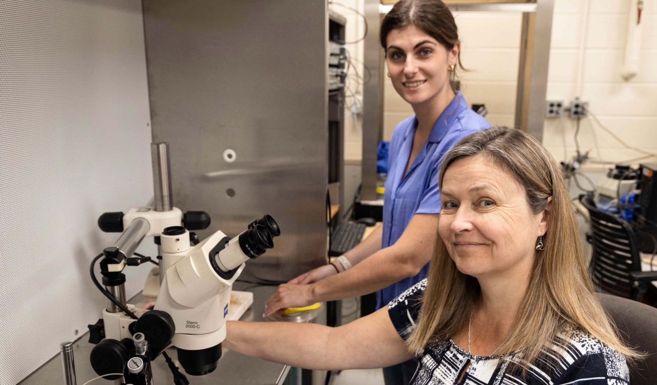 A smiling woman sits in front of a microscope with a woman standing behind her in a laboratory setting.