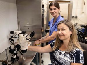 A smiling woman sits in front of a microscope with a woman standing behind her in a laboratory setting.