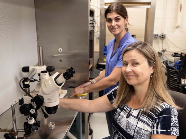 A smiling woman sits in front of a microscope with a woman standing behind her in a laboratory setting.