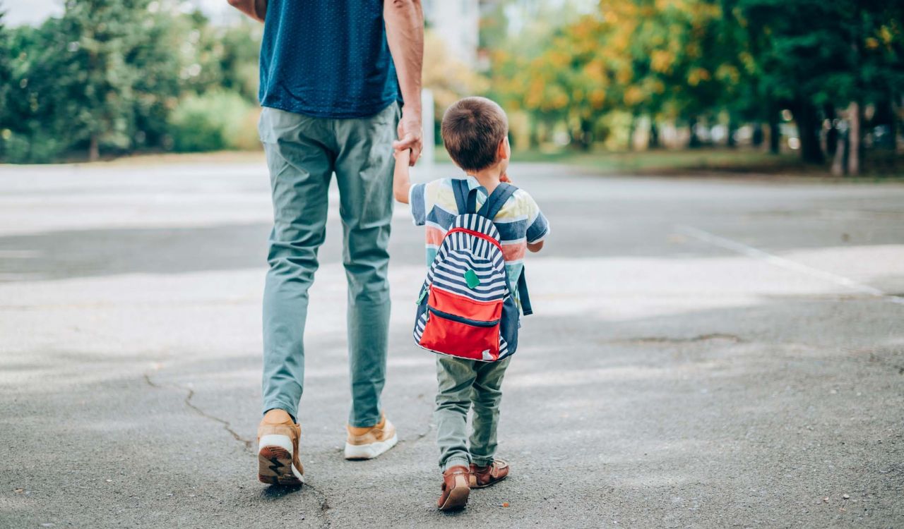 A parent holds the hand of a child wearing a backpack as they walk forward together.