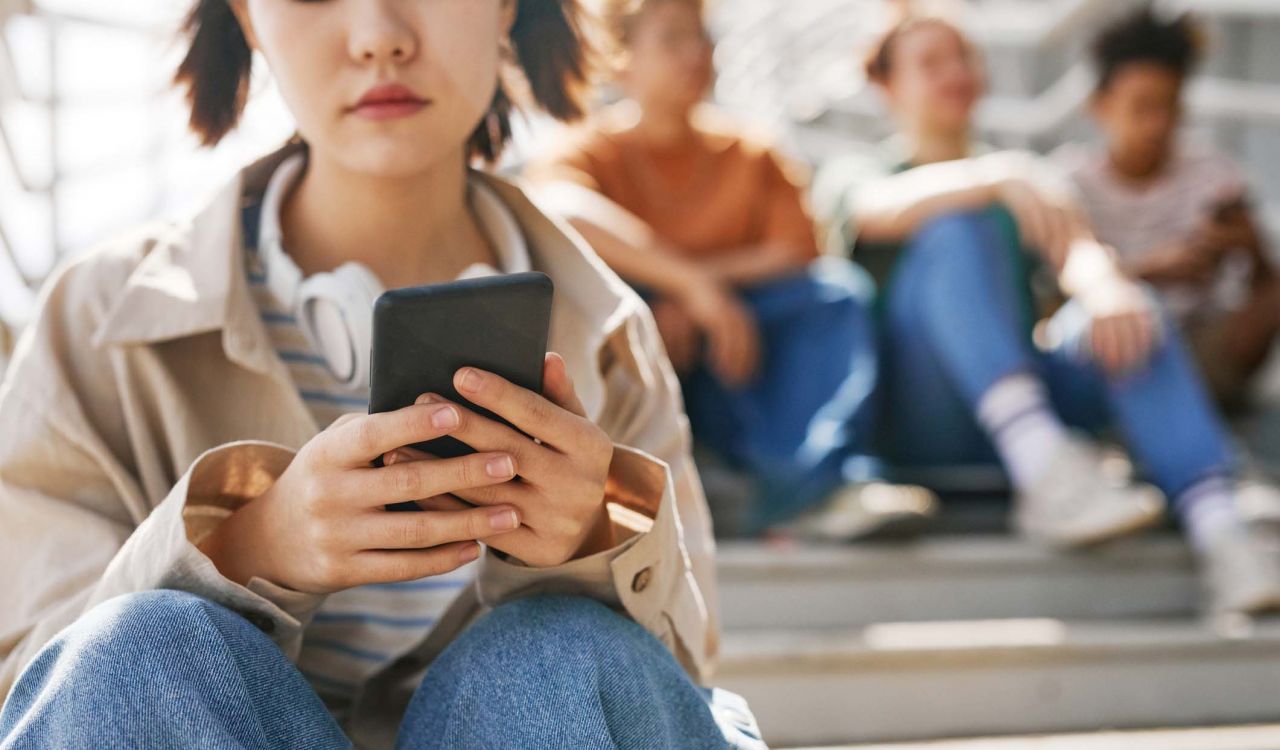 A teenage girl sits alone using her cellphone with another group of teenagers sitting in the background.