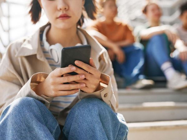 A teenage girl sits alone using her cellphone with another group of teenagers sitting in the background.
