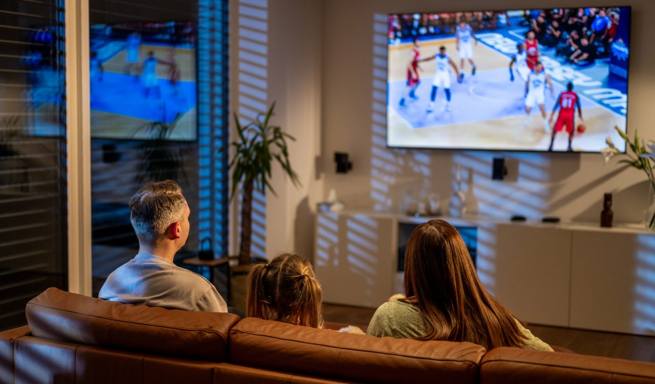 Two parents and a child, seen from behind, watch a basketball game on TV while sitting on a sofa.