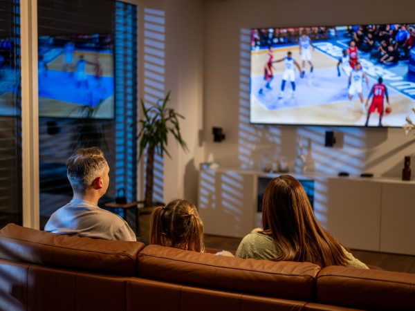 Two parents and a child, seen from behind, watch a basketball game on TV while sitting on a sofa.