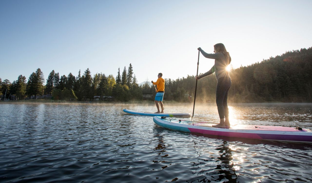 Couple paddle stand up paddleboards on tranquil mountain lake at sunrise, mist rises from Lost Lake, forest and mountains, Whistler, B.C.