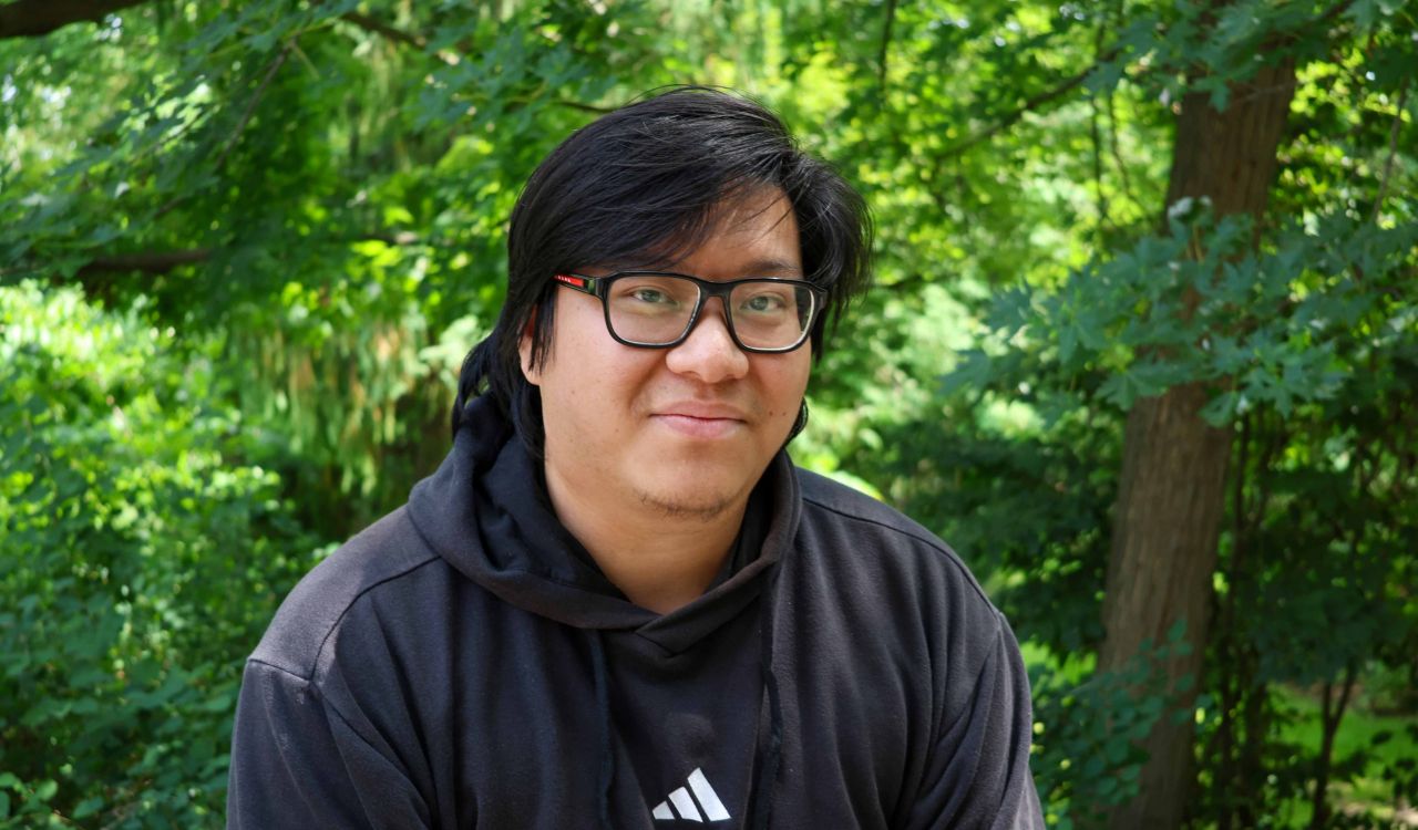 Steven Duong smiles while sitting outside in front of green trees.