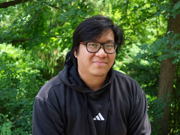 Steven Duong smiles while sitting outside in front of green trees.