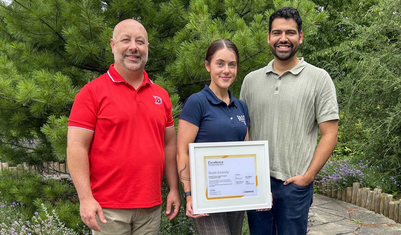 Three people pose for a photo in a garden with a large framed certificate.