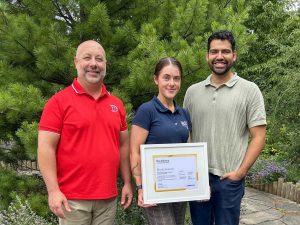 Three people pose for a photo in a garden with a large framed certificate.