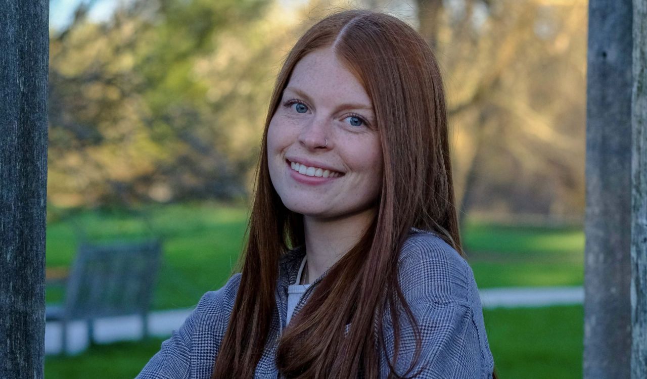 A young woman smiles for a photo with a green space and trees behind her.