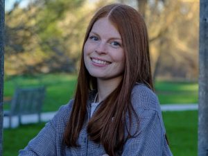 A young woman smiles for a photo with a green space and trees behind her.
