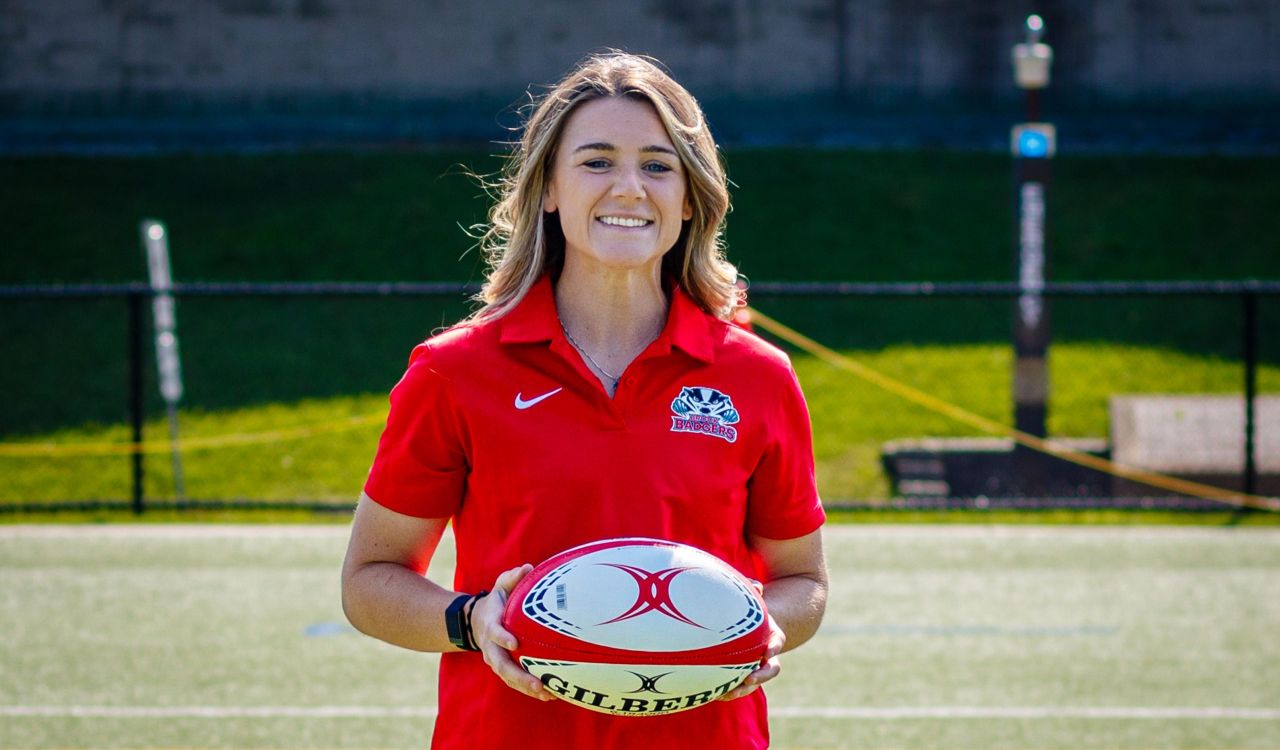 A woman in a red shirt poses with a rugby ball on a field.