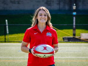 A woman in a red shirt poses with a rugby ball on a field.