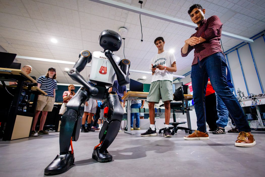 A small humanoid robot stands in the foreground of a robotics lab while a group of students watches. One student holds a controller as others observe the robot's movements during a demonstration.