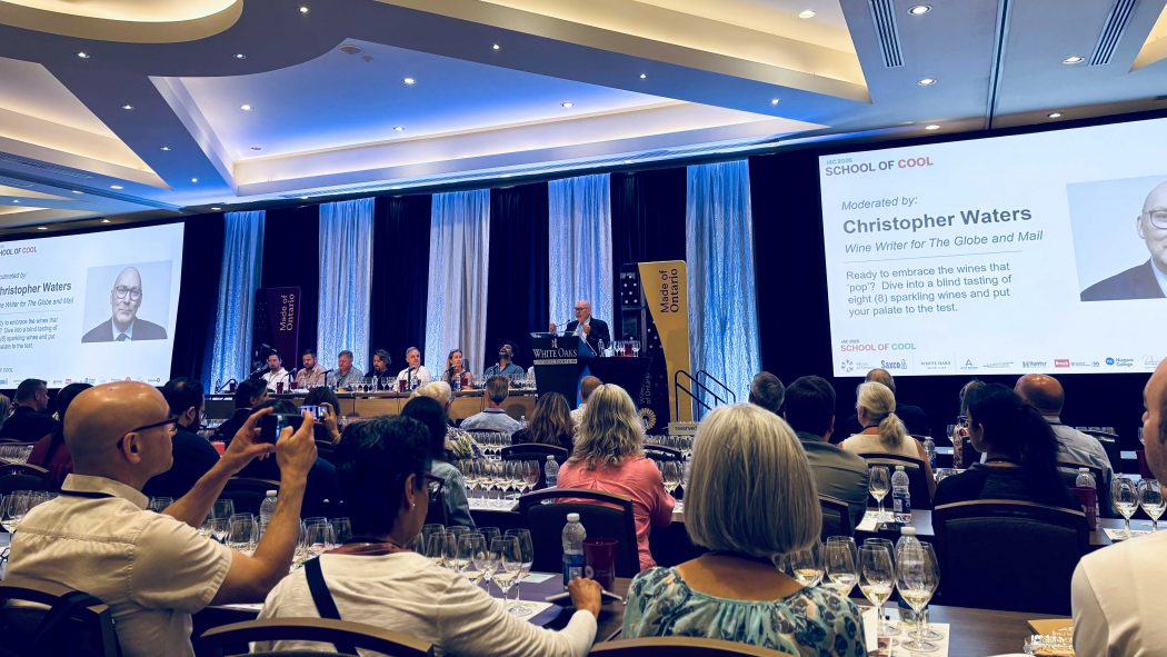 A large group of wine enthusiasts sit in a conference room at tables set with wine glasses listening to presenter Chris Waters speak about sparkling wine.