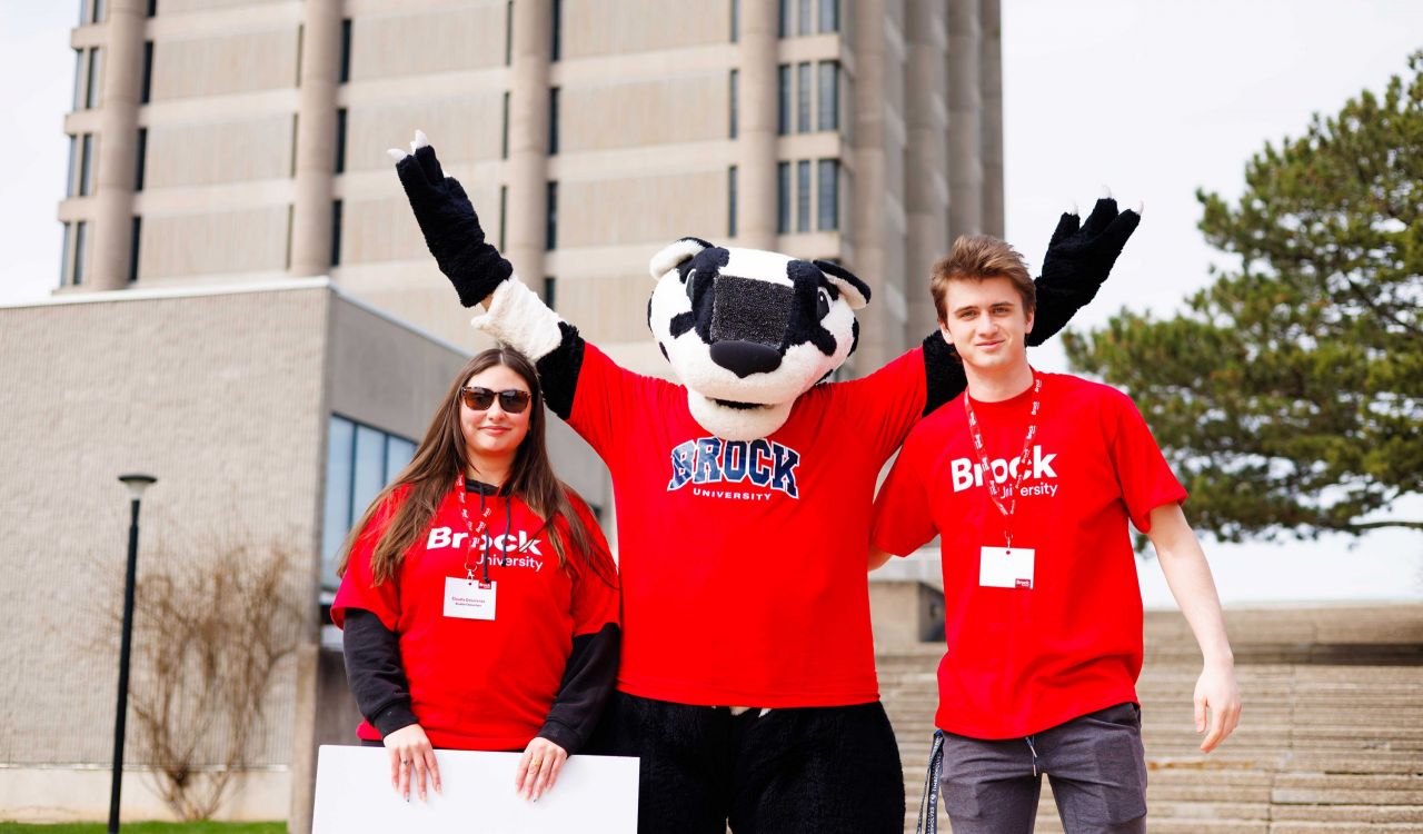 Two people in red Brock University-branded shirts stand on either side of Boomer the Badger, a large badger mascot.