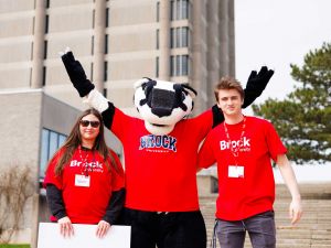 Two people in red Brock University-branded shirts stand on either side of Boomer the Badger, a large badger mascot.