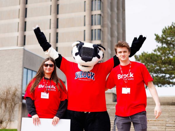 Two people in red Brock University-branded shirts stand on either side of Boomer the Badger, a large badger mascot.