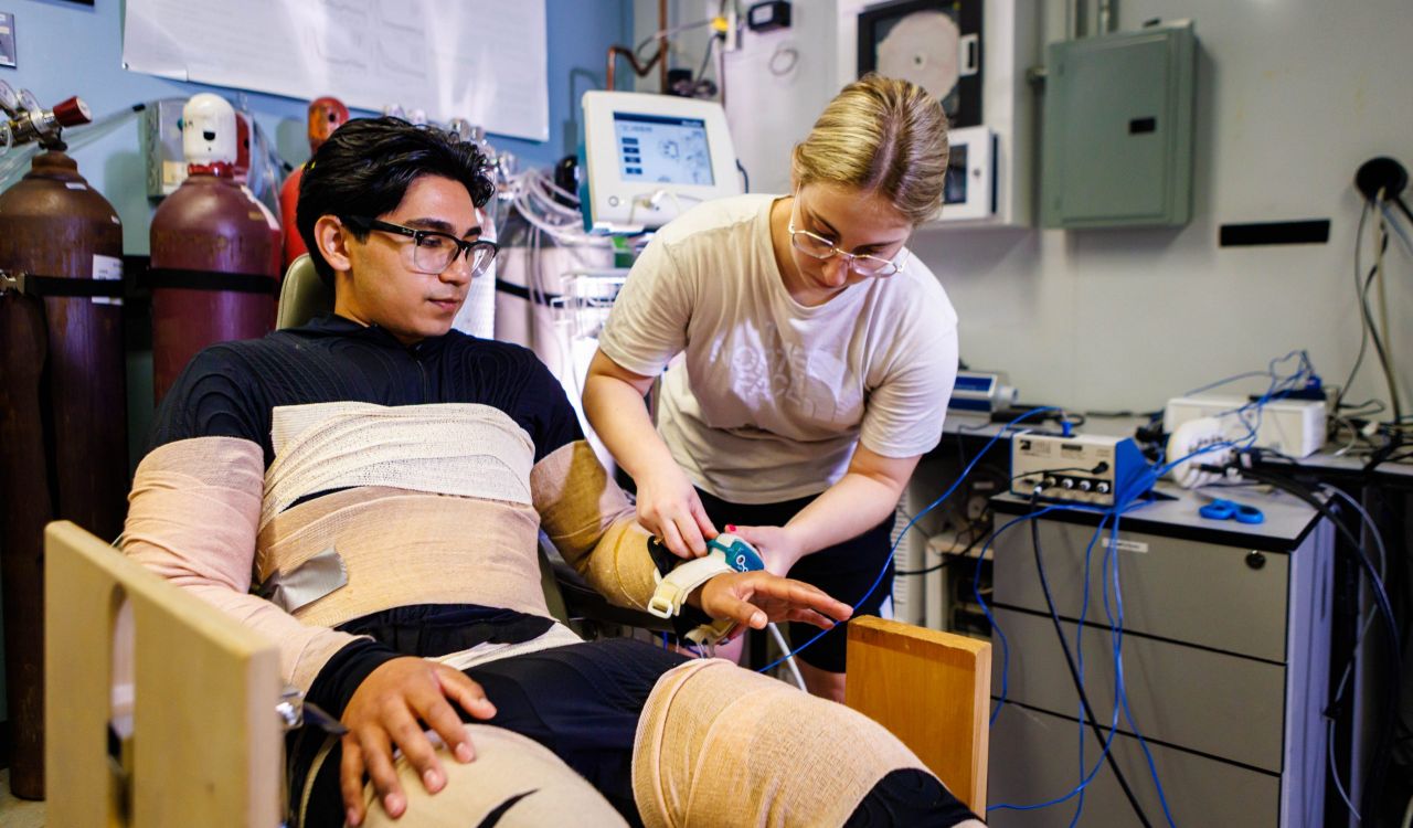 A woman places a device on the wrist of a man who is wrapped in bandages and seated in a chair as part of a research experiment.