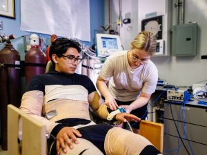 A woman places a device on the wrist of a man who is wrapped in bandages and seated in a chair as part of a research experiment.
