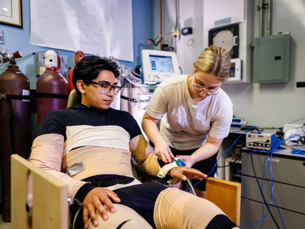 A woman places a device on the wrist of a man who is wrapped in bandages and seated in a chair as part of a research experiment.