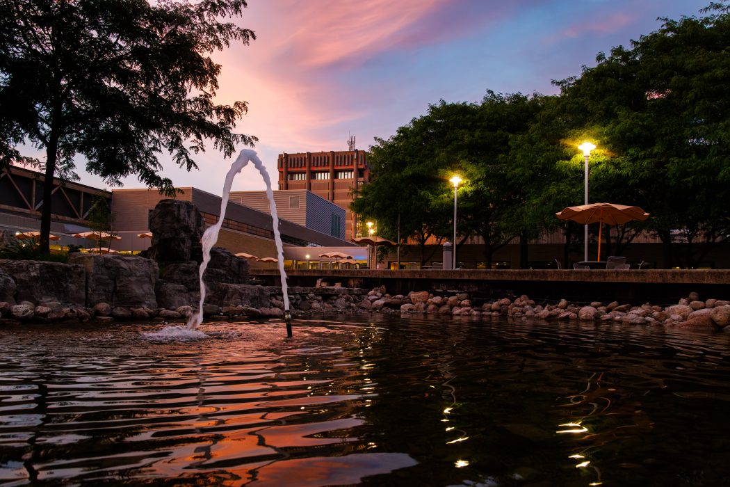 A newly installed fountain sprays water into the restored pond in Brock University’s Walker Complex courtyard at sunset, with trees, outdoor seating and Schmon Tower visible in the background.