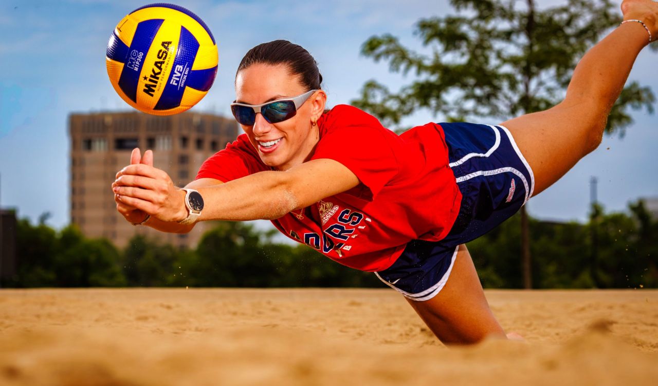 Close-up of a smiling woman diving into the sand, extending her arms to strike a volleyball, with the Brock tower in the background.
