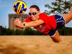 Close-up of a smiling woman diving into the sand, extending her arms to strike a volleyball, with the Brock tower in the background.