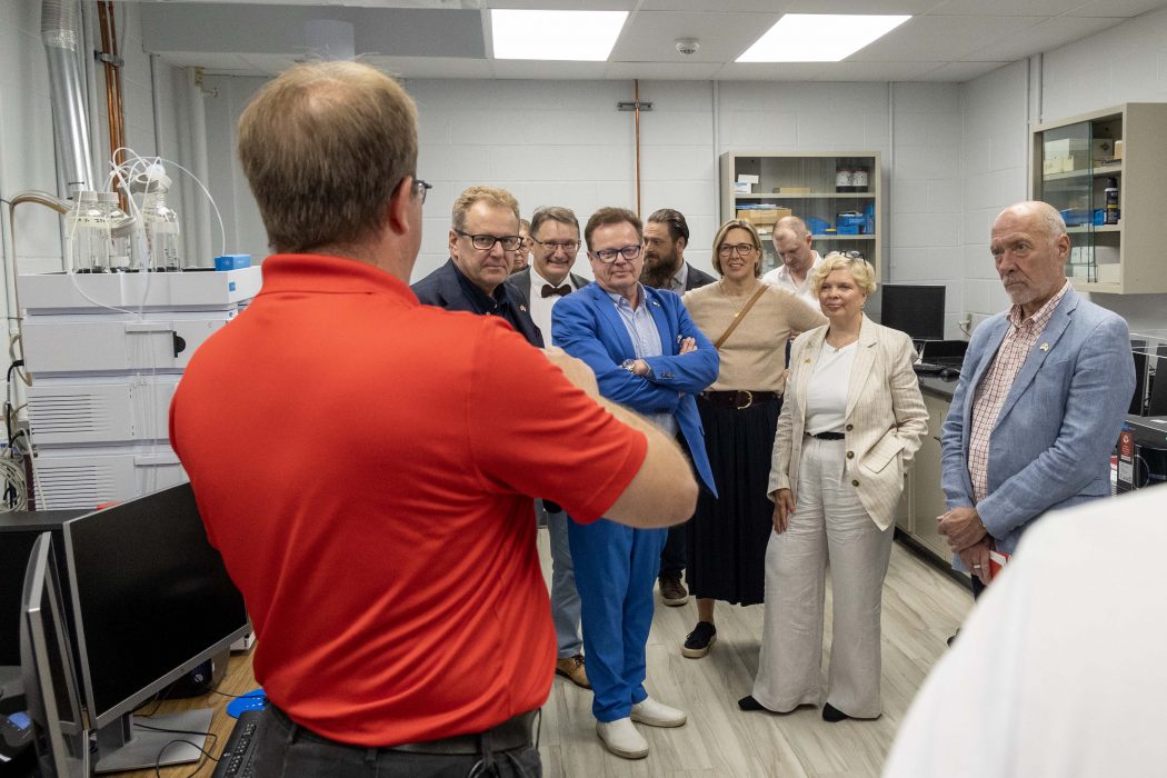 A man in a red shirt speaks to a group of business people during a tour of a lab space.