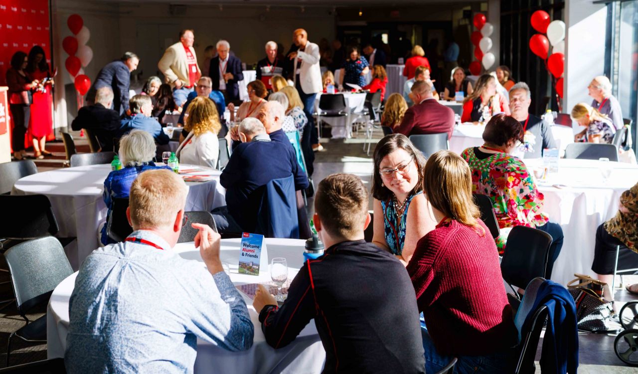 A room full of people sit at table or stand and chat at a university reunion event.
