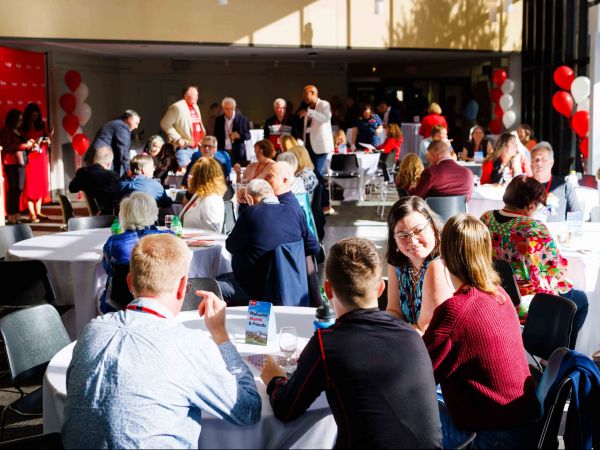 A room full of people sit at table or stand and chat at a university reunion event.