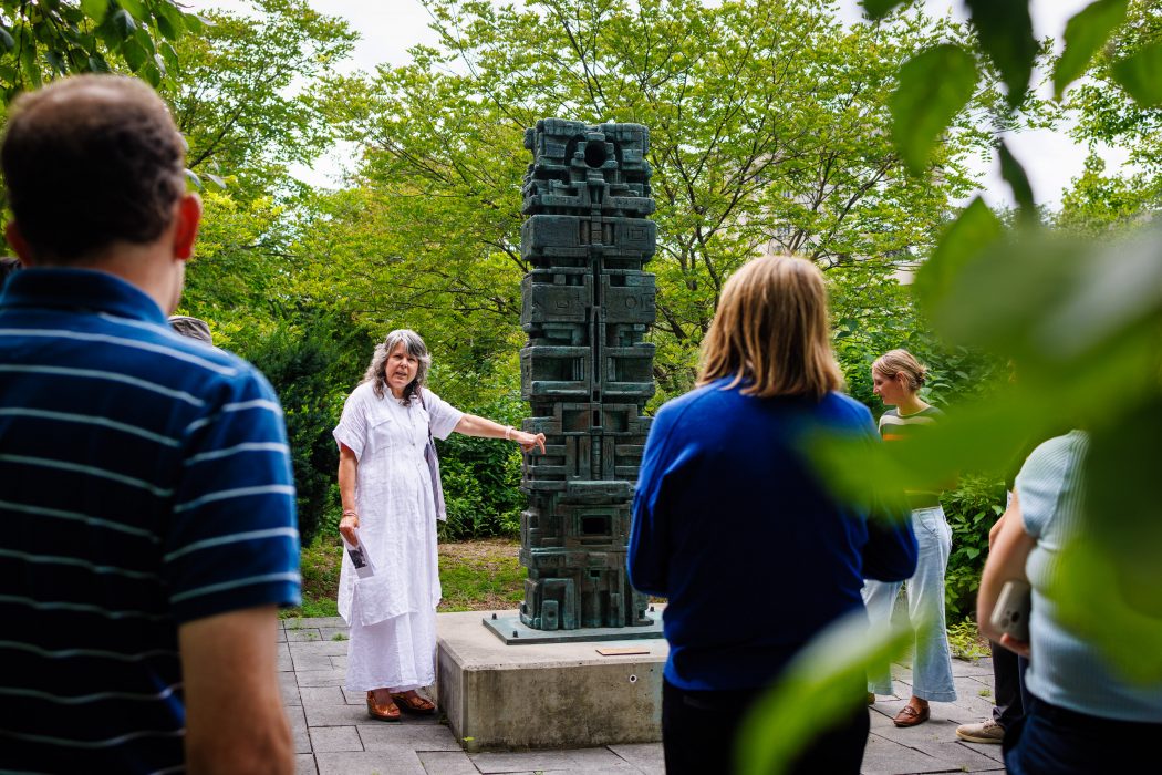A woman points at a sculpture in a garden while guiding a group of tour participants.