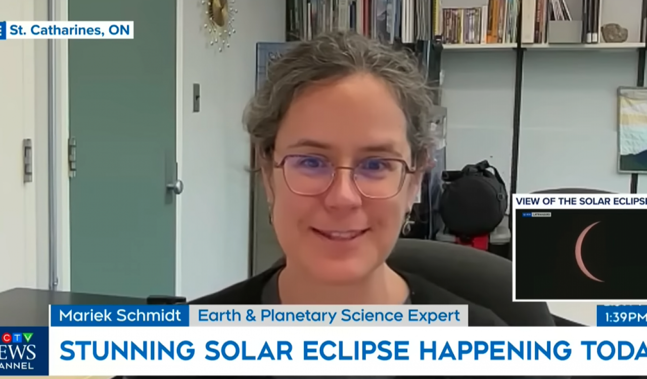 A woman is seated at a desk in an office looking straight ahead. A small inset box shows a view of the solar eclipse, only a thin crescent of the sun is visible behind the moon.