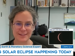 A woman is seated at a desk in an office looking straight ahead. A small inset box shows a view of the solar eclipse, only a thin crescent of the sun is visible behind the moon.