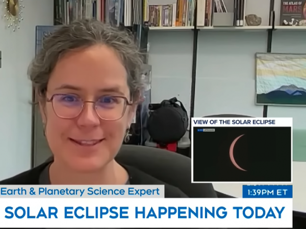 A woman is seated at a desk in an office looking straight ahead. A small inset box shows a view of the solar eclipse, only a thin crescent of the sun is visible behind the moon.