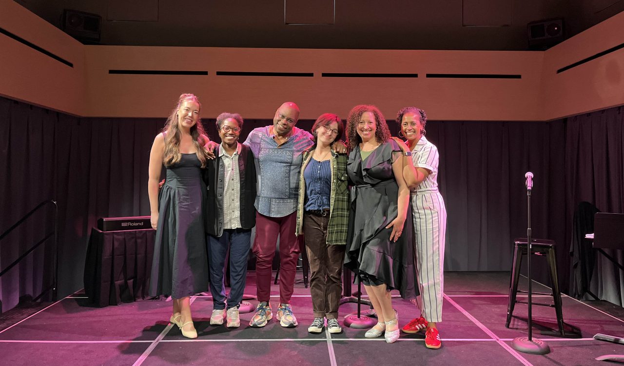 A group of people stand together shoulder to shoulder on a small black box professional theatre stage.