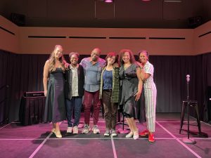 A group of people stand together shoulder to shoulder on a small black box professional theatre stage.