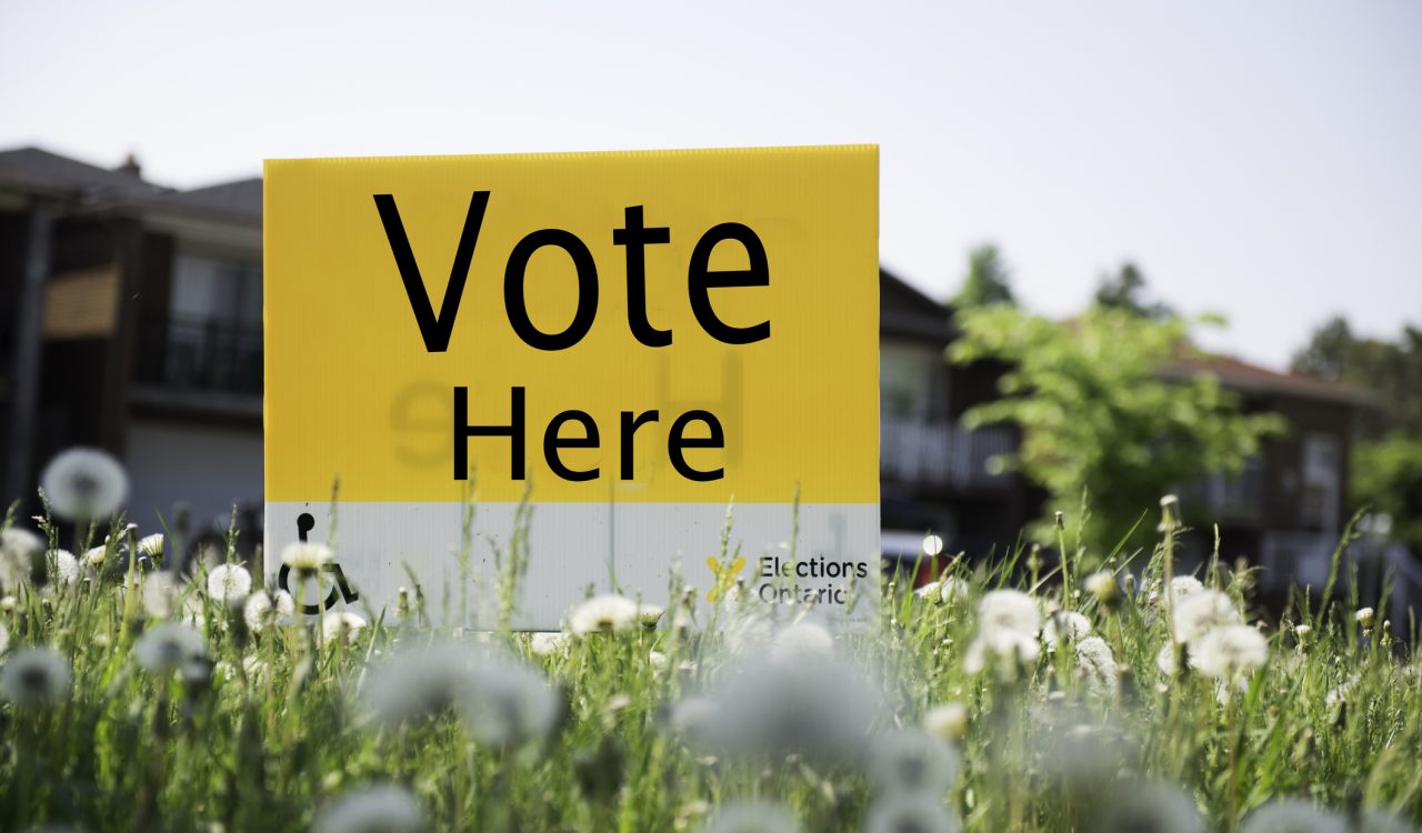 A yellow and white "Vote Here" sign in the grass outside a building.