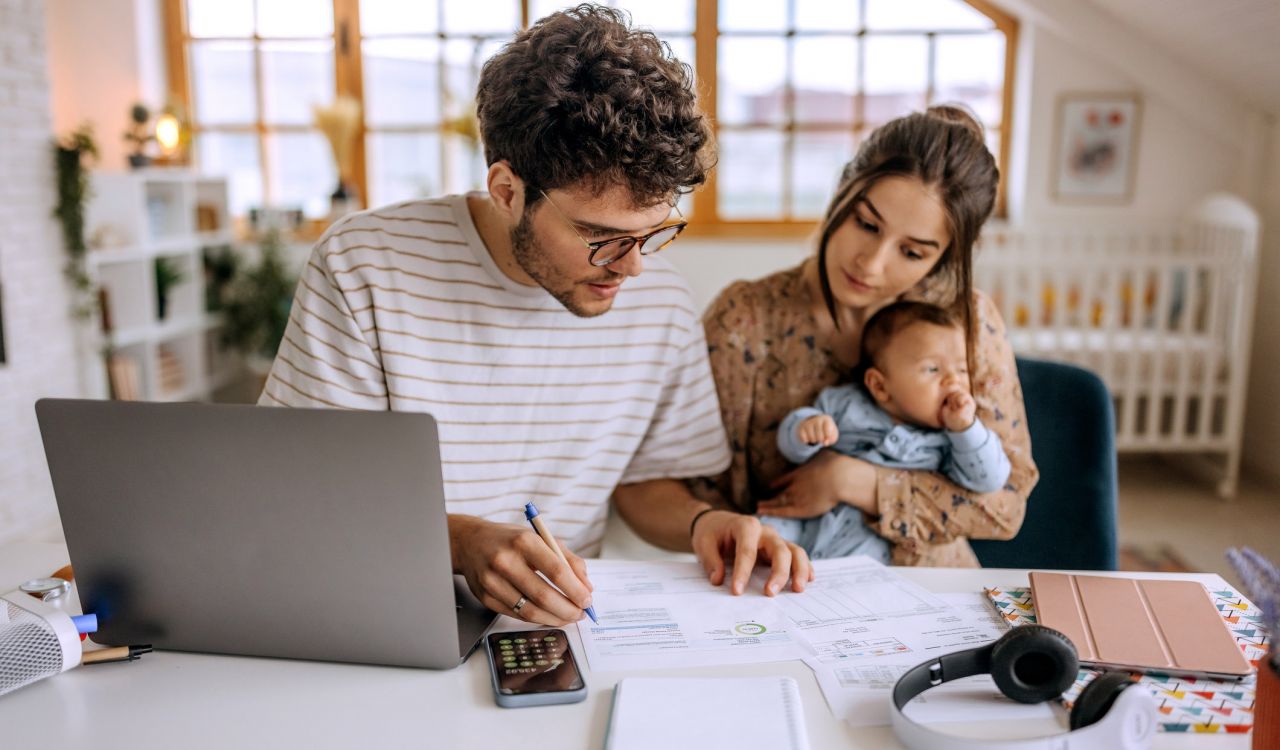 Two young adults look at financial papers while sitting a table with their baby.