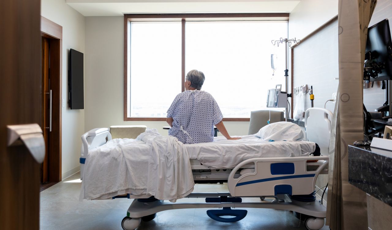An elderly person wearing a hospital gown sits on a hospital bed looking around a hospital room.