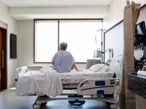 An elderly person wearing a hospital gown sits on a hospital bed looking around a hospital room.
