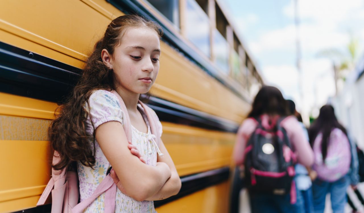A sad young girl stands beside a yellow school bus while other students get onto the bus.