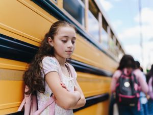 A sad young girl stands beside a yellow school bus while other students get onto the bus.
