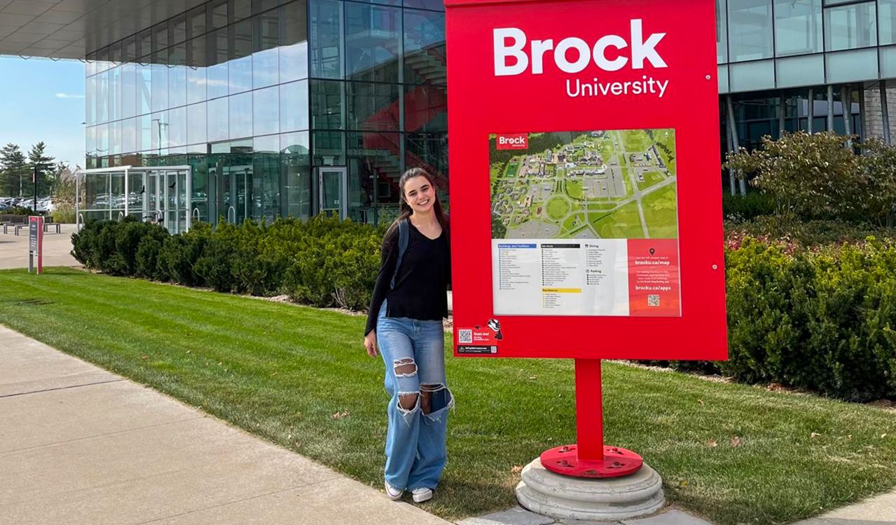 A young woman stands beside a red sign and map outside a Brock University building.