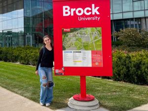 A young woman stands beside a red sign and map outside a Brock University building.