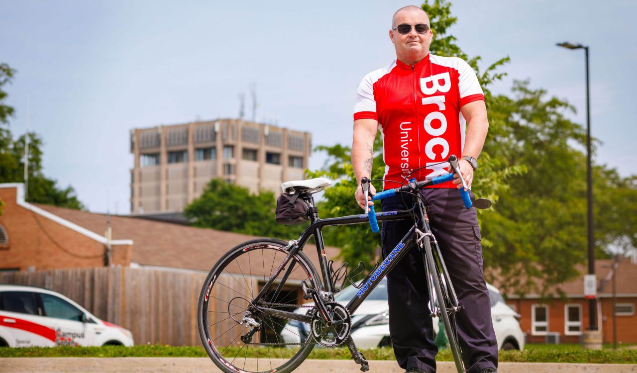 A man stands behind his bicycle on a campus road, with a blue sky, greenery and the Schmon Tower in the background.