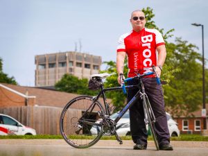 A man stands behind his bicycle on a campus road, with a blue sky, greenery and the Schmon Tower in the background.