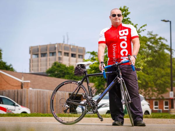A man stands behind his bicycle on a campus road, with a blue sky, greenery and the Schmon Tower in the background.