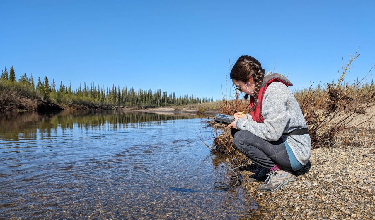 A woman holding, and looking at, a device crouches down next to a small creek with a rugged shoreline in the background.